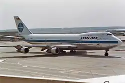 Photo d'un avion de Pan Am vu de trois quart face sur le tarmac gris d'un aéroport. L'avion, en gros plan, est stationné et entouré de bâtiments de faible hauteur, d'un autre avion et d'une forêt en arrière-plan.