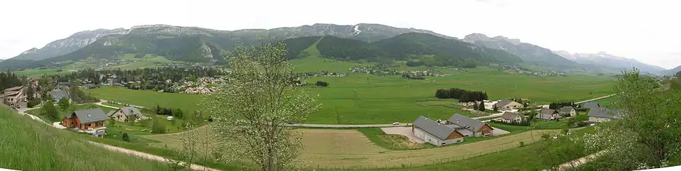 Un plateau verdoyant parsemé de maisons au premier plan, avec des crêtes montagneuses en arrière-plan