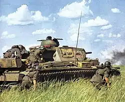 Photo en couleur montrant un char dans de la végétation. Tois soldats se protège derrière sa tourelle, tandis que deux solats marchent courbés à côté. On les voit de dos.