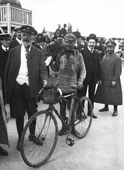 Photographie en noir et blanc montrant un cycliste couvert de boue à l'arrivée d'une course, entouré de plusieurs personnes.