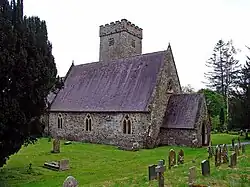 Photo d'un bâtiment au toit d'ardoises très pentu avec une tour crénelée au milieu, entouré d'une pelouse verte jonchée de pierres tombales