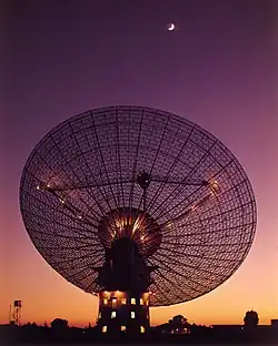 Photographie d'une antenne parabolique pointant vers le ciel, durant le crépuscule, la lune (croissant) étant visible sur le haut de l'image.