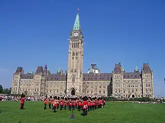 Photo en couleurs de militaires en uniformes rouges sur une place devant le bâtiment du Parlement du Canada