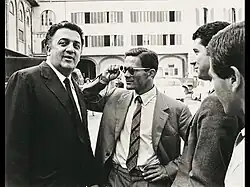 Quatre hommes, dont un avec des lunettes, sur un parking - sur la gauche, on reconnaît Federico Fellini et Pier Paolo Pasolini