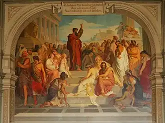 Paul, habillé en rouge, lève le bras en haut de marches. La foule est rassemblée autour de lui, à son niveau ou sur les marches.