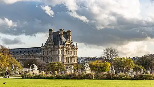 Sous ciel gris, un bâtiment classique couronne un jardin de pelouses vertes, bosquets et statues.