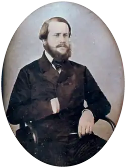 Photographie d'un jeune homme barbu assis dans un fauteuil avec sa main droite rentrée dans l'avant de sa veste à double boutonnage