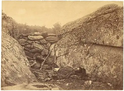 Photographie en noir et blanc d'un soldat mort, allongé sur le dos, légèrement tourné vers l'objectif, au milieu de rochers