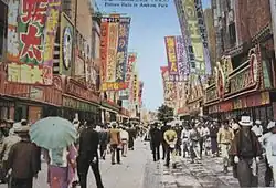 Photo colorisée d'une rue japonaise des années 1930, avec beaucoup de passants et de nombreuses banderoles accrochées aux façade.