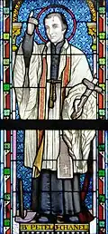 Vitrail en couleurs montrant un homme blanc habillé en blanc (chasuble), levant son bras et son index droit et tenant une croix dans la main gauche, sa tête surmontée d'une auréole rouge sur fond bleu. Le texte en bas du personnage indique "B. Petelo Chanel" (bienheureux Pierre Chanel).