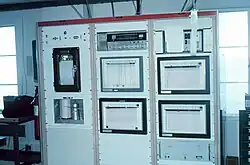 Dans un bureau, une large armoire industrielle métallique, haute d'environ deux mètres, composée de trois panneaux verticaux, lesquels comportent des appareils scientifiques et des enregistreurs graphiques.