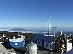 Depuis une hauteur, plusieurs bâtiments et cabanons de plain-pied ; le bâtiment de droite possède plusieurs mâts et antennes sur son toit, maintenus par des haubans. À l'arrière-plan, au loin, une montagne, l'océan et le ciel.