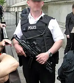 Agent de police en uniforme portant un gilet pare-balles réglementaire et un fusil d'assaut, dans une rue.