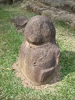 Une petite statuette en pierre avec une tête ronde complètement lisse et un corps rond et trapu. On voit la figurine de face sur sa gauche, et le bras gauche est levé contre le côté du corps. Le bras descend de l'épaule et est plié vers l'avant au coude, horizontal jusqu'au poignet, qui est plié vers le bas, avec les doigts représentés par des rainures sculptées. La statuette n'a pas de jambes, et repose sur un coin d'herbe devant des marches en pierre.