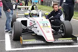 Photographie d'une Formule 4 blanche et jaune, vue de face, à l'arrêt sur une grille de départ. Le pilote est dans la voiture, entouré de deux mécaniciens.