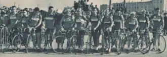 Photo en noir et blanc d'une douzaine d'hommes en tenue de cycliste, debout à côté de leurs vélos qui se tiennent les uns à côté des autres.