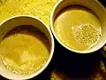 Deux verres de mélange de pinol dans du lait chaud, mélangés de manière presque homogène. Les restes de poudre de pinol flottent à la surface du lait.