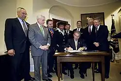 Photographie en couleurs d'une dizaine de personnes dans un bureau. Un homme est assis et signe un document sur une table, les autres le regardent, debout autour de lui.