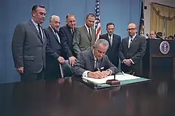 Un homme, assis, signant un document, six hommes, debout, derrière lui.