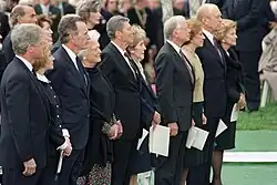 Une rangée de 10 hommes et femmes en couples se tiennent debout lors d'une cérémonie, une foule est présente à l'arrière-plan