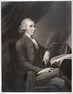 Portrait d’un homme assis sur une chaise et accoudé à une table couverte de livres et publications.