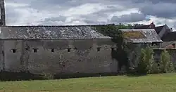 Photographies en couleurs d'un bâtiment oblong couvert d'une toiture en ardoise et percé de petites baies carrées, une étendue herbeuse le longeant au premier plan.