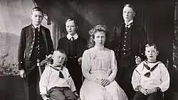Photographie en noir et blanc montrant six enfants et adolescents : trois garçons en costume noir, deux garçons en uniforme de marin et une fille en robe blanche.