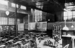 Image en noir et blanc d'une grande pièce avec des chaises devant un bureau et des tableaux. Les fenêtres sont munies de barreaux.