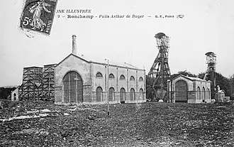 Photo noir et blanc d'un ensemble de bâtiments industriels années 1900 avec deux grands chevalements (tour avec bigue) métalliques et cheminée d'usine.