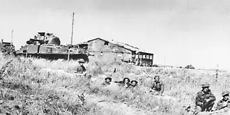 Photographie en noir et blanc d'un char militaire avec des soldats dans un champ devant un bâtiment en ruine