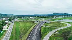 Vue aérienne d'un échangeur en trompette emprunté par une voie rapide avec deux voies de chaque côté. En second plan, on distingue le village de Maïma avec de nombreuses maisons. Des montagnes de faible altitude se révèlent en arrière-plan.