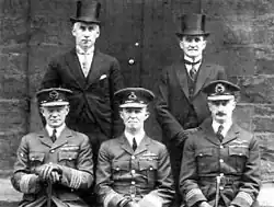 Portrait de cinq hommes, trois assis portant des uniformes militaires sombres avec des casquettes à visière, et deux debout derrière portant des costumes formels avec des chapeaux hauts de forme.