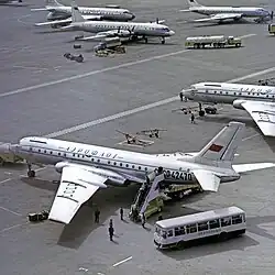 tarmac d'un aéroport. au premier plan, des passagers embarquent dans un Tu-104, depuis un bus. en arrière plan, un autre tu104 et un il-18 à hélices.
