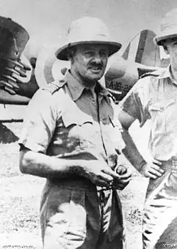 Portrait d'un homme en uniforme militaire tropical. Il porte un chapeau à godets et l'on aperçoit des avions militaires à l'arrière-plan.