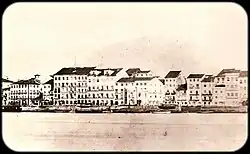 Une vieille photo montrant des tas de matériaux de construction sur la berge d'un fleuve avec de grands bâtiments blancs qui bordent la rive opposée