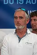 Vue en couleur du buste d'un homme en polo blanc, barbe et cheveux gris, des lunettes de soleil sur la tête.