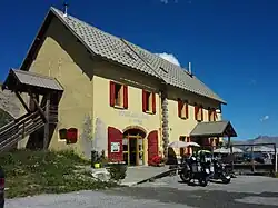 Façade et pignon d’une maison à un étage, murs jaunes et volets rouges.