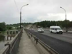 Photographie prise sur le trottoir d'un pont (sur la rivière Inia), avec des véhicules circulant dans la direction du photographe. Les véhicules dans l'autre sens sont sur un autre pont parallèle en arrière-plan. Au fond se trouve une colline recouverte d'arbres.