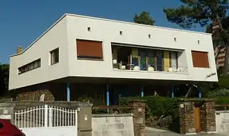Photographie d'une villa parallélépipédique blanche, percée d'une grande loggia rectangulaire et de plus petites fenêtres aux huisseries de bois vernis. Le rez-de-chaussée est constitué d'un soubassement, recouvert de pierres sombres, et d'une partie évidée soutenue par de fines colonnettes bleues