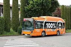Photographie en couleurs d’un autobus électrique orange en essais à Rumilly, roulant devant une haie d'arbres.
