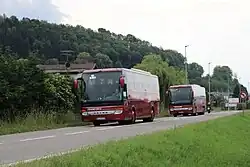 Photographie en couleurs de deux autocars rouges sur une route bordée par un pré vert. En arrière-plan se détachent une haie et le toit de quelques habitations.