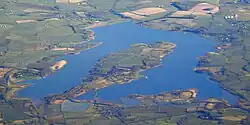 Photo aérienne d'une étendue d'eau entourée de champs verts et de petits villages. Un îlot de forme ovale se dresse au milieu de l'eau, relié à la terre ferme par un isthme en bas de l'image
