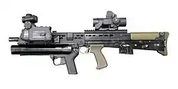 photographie en couleur montrant de profil un fusil d’assaut avec plusieurs modules fixés autour et sous le canon.