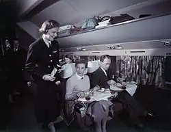 passagers installés, une hôtesse de l'air en uniforme noir leur présente une bouteille de vin.