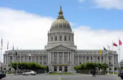 Un grand bâtiment sur trois étages avec une aile de chaque côté d'une tour imposante. Le bâtiment est blanc, avec un dome or et bleu au sommet de la tour. Devant le bâtiment on trouve une place encadrée de mâts pour drapeau et d'arbres.
