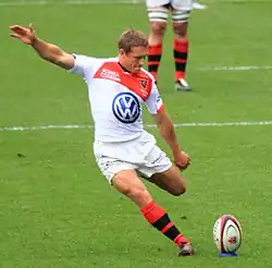 Wilkinson vêtu de blanc, dans le mouvement du tir, jambe gauche repliée, bas droit tendu légèrement vers le haut.
