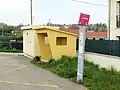 Poteau d'arrêt aux couleurs actuelles du réseau, avec un abribus en béton à côté.