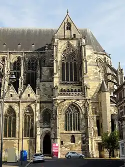 Petit transept avec sa rosace en forme de fleurs et la grande fenêtre des parties basses. À gauche, le portail Lamoureux de la basilique, fermé par une grille en fer forgé.