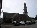 L'église Saint-Cornély (au premier plan la place de l'église et ses voitures garées, au second plan l'église avec son clocher à droite).