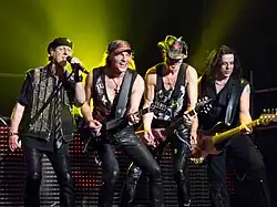 Photo de quatre rockeurs sur une scène, habillés en cuir noir. Celui de gauche chante dans un micro et les trois autres jouent de la guitare ou de la basse électriques. Derrière eux, on voit un halo de lumière verte et un décor fait d'ampoules rouges.